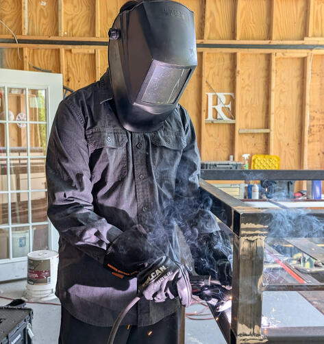 Man welding a metal frame for a custom LED sign inside a workshop.